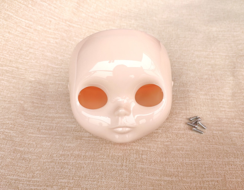 Puede incluir: Una cabeza de mu&ntilde;eca de color rosa p&aacute;lido con grandes cuencas oculares y una boca peque&ntilde;a y cerrada. La cabeza es lisa y brillante, con unos peque&ntilde;os tornillos a la derecha. La cabeza de la mu&ntilde;eca es probablemente para un proyecto de mu&ntilde;eca personalizado.