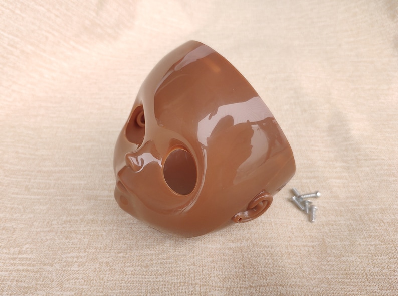 Puede incluir: Una cabeza de mu&ntilde;eca marr&oacute;n con acabado brillante, cortada por la mitad. La cabeza presenta cuencas de los ojos, nariz, boca y detalles de las orejas. Varios tornillos plateados peque&ntilde;os est&aacute;n junto a la cabeza.