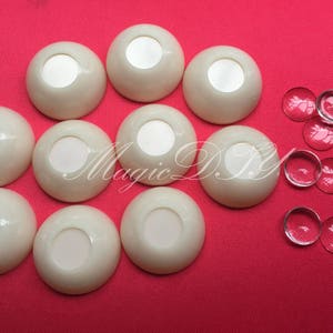 May include: Ten white plastic eye sockets, each with a small, round, white centre, arranged in a circle on a red surface.  There are also several clear glass beads scattered around the eye sockets.