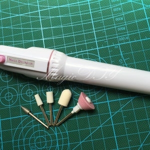 Peut inclure: Un appareil électrique blanc pour le soin des ongles avec un bouton rose et le texte "Nails Decorator" sur le côté. L'appareil est livré avec cinq accessoires différents, dont un cône de ponçage rose, un cône de ponçage blanc et trois mèches métalliques.