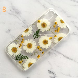 May include: A clear phone case with a pressed flower design. The case features white daisies with yellow centers and green leaves.