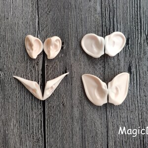 May include: Four pairs of elf ears made of a light beige colored material. The ears are in various shapes and sizes. The ears are on a dark wood background.