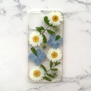 May include: A clear phone case with pressed flowers and leaves. The flowers are white with yellow centers and the leaves are green. There are also blue flowers in the design.