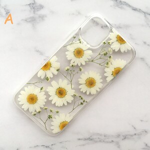 May include: A clear phone case with a pressed flower design. The case features white daisies and small white flowers pressed into the clear plastic.