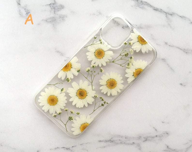 May include: A clear phone case with a pressed flower design. The case features white daisies and small white flowers pressed into the clear plastic.