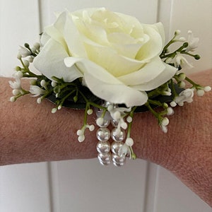 May include: A white rose wrist corsage with white pearl accents and greenery.