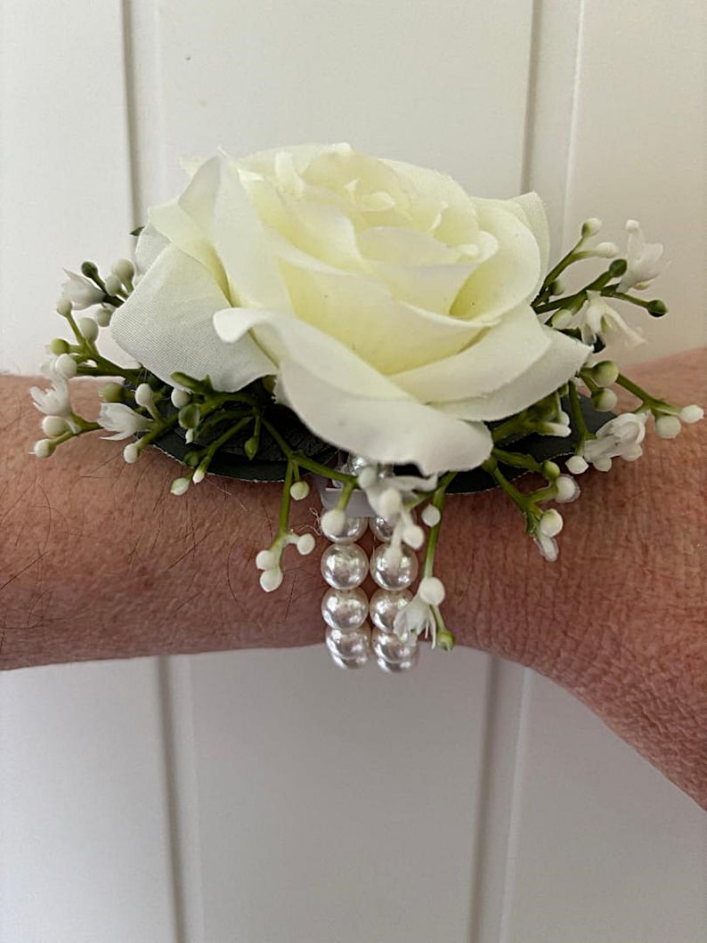 May include: A white rose wrist corsage with white pearl accents and greenery.