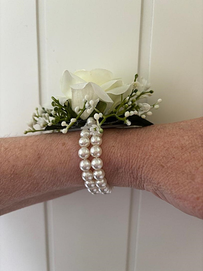 May include: A white rose with green leaves and white pearl beads on a wrist.