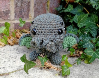 May include: A crocheted plush toy resembling a creature with large eyes and green appendages. The toy is gray and green and is sitting on a concrete surface with green ivy.