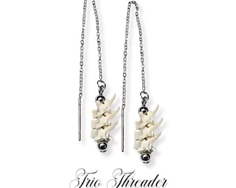 Puede incluir: Pendientes de hilo con una cadena y un grupo de pequeñas vértebras blanquecinas. Los pendientes tienen una bola plateada en la parte inferior y están suspendidos de una cadena. El texto "Trio Threader" y "Rattlesnake Vertebrae Threader Earrings" está en la parte inferior.