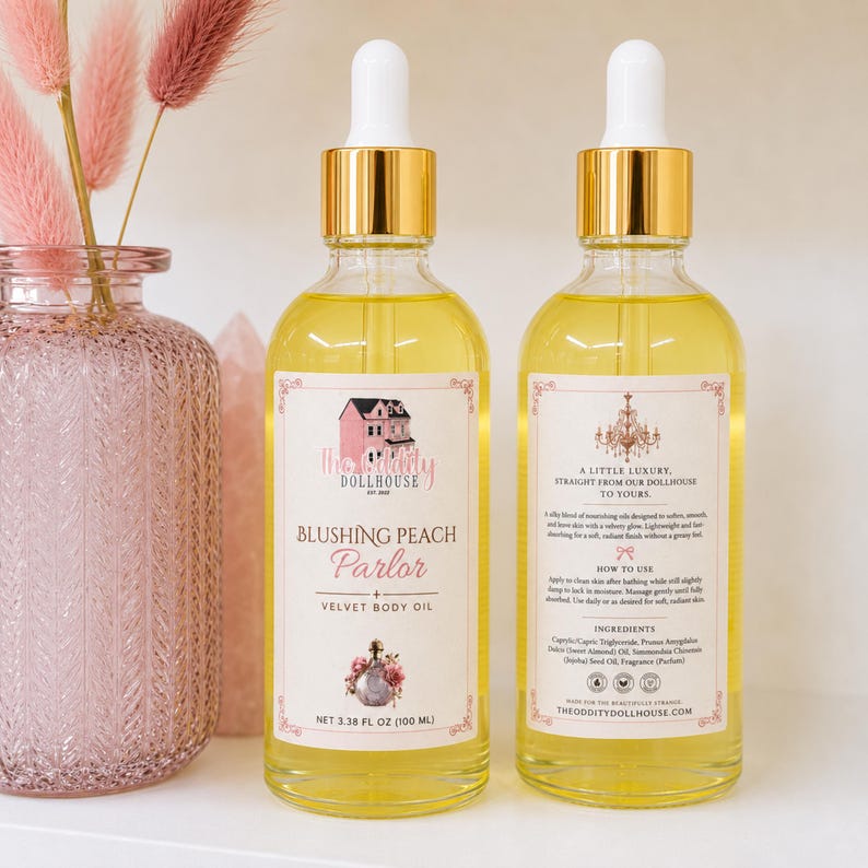 May include: Two clear glass bottles of Blushing Peach Parlor Velvet Body Oil with gold caps and white droppers. The front label features a pink dollhouse illustration and text, while the back label lists ingredients and instructions in 3.38 fl oz.
