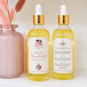 May include: Two clear glass bottles of Velvet Bombshell body oil with gold dropper caps. Each bottle contains 3.38 fl oz of yellow liquid. The front label features a pink house illustration and text, while the back label lists ingredients.