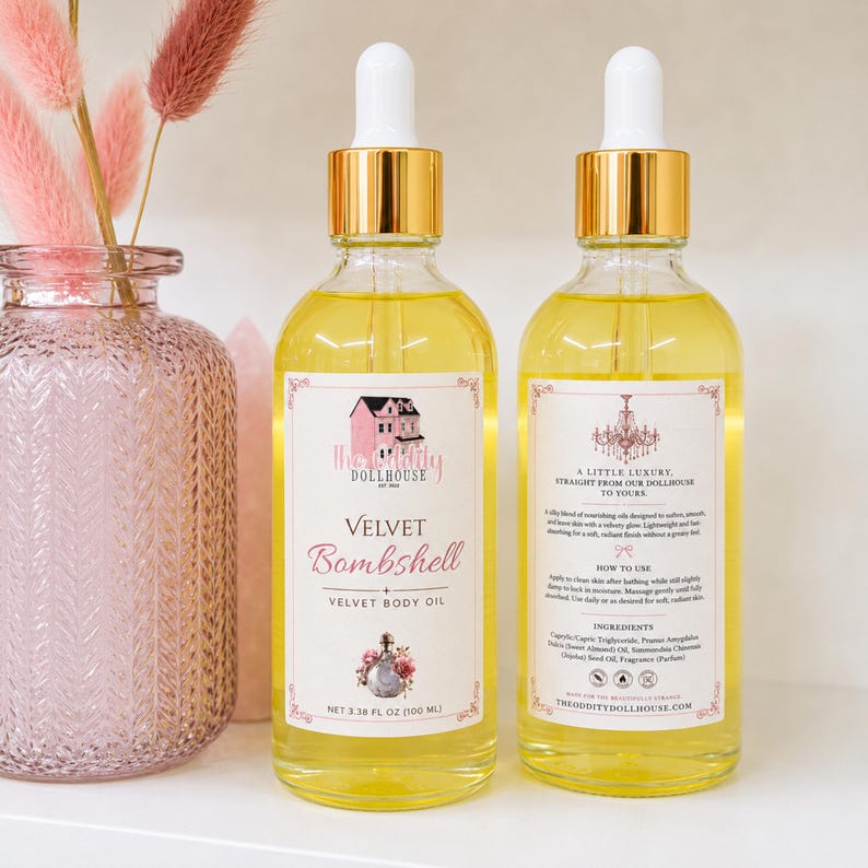 May include: Two clear glass bottles of Velvet Bombshell body oil with gold dropper caps. Each bottle contains 3.38 fl oz of yellow liquid. The front label features a pink house illustration and text, while the back label lists ingredients.