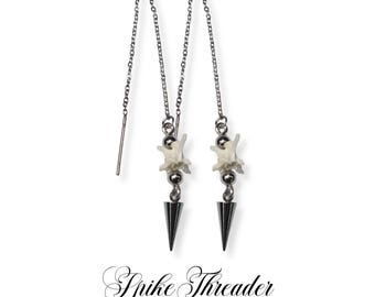 May include: Silver threader earrings with a unique design. Each earring features a small, white bone, a silver bead, and a black, cone-shaped spike. The earrings are suspended from a delicate silver chain. The text "Spike Threader" and "Rattlesnake Vertebrae Threader Earrings" are at the bottom.