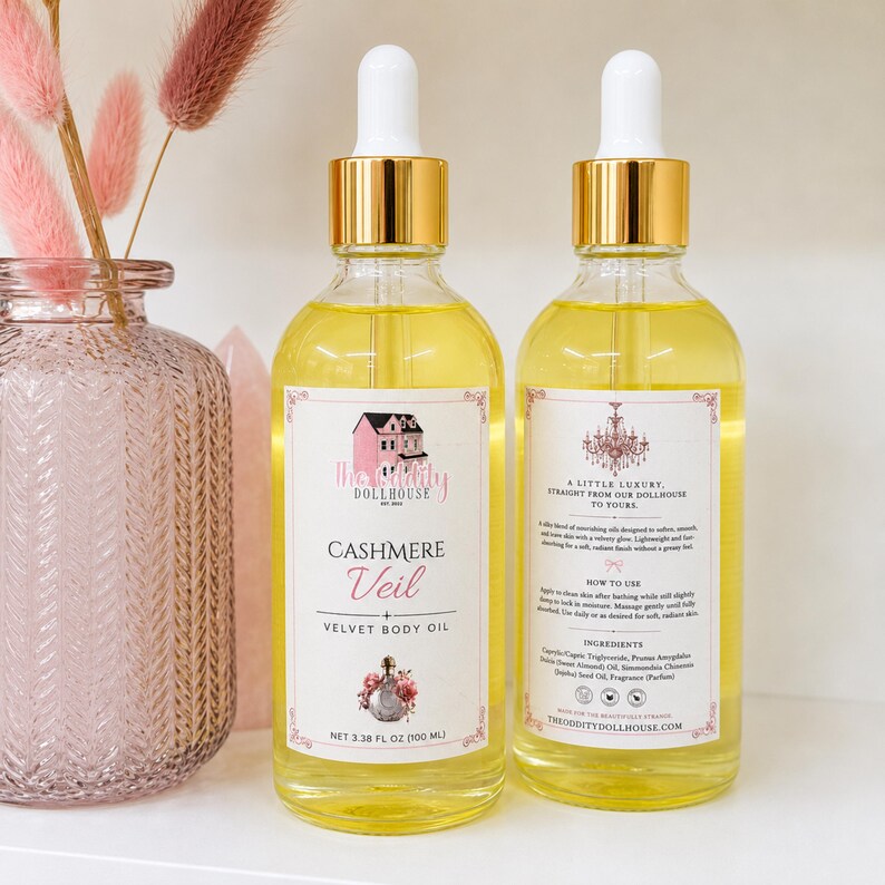May include: Two clear glass bottles of Cashmere Veil velvet body oil with gold caps and white droppers. Each bottle features a white label with black text, a pink dollhouse illustration, and a net volume of 3.38 fl oz (100 ml) printed on the front.