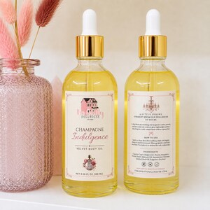 May include: Two clear glass dropper bottles filled with yellow body oil, featuring gold caps and white labels. The front label reads Champagne Indulgence Velvet Body Oil, net 3.38 fl oz (100 ml). The back label includes instructions and ingredients.