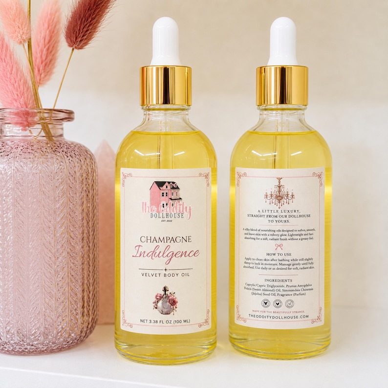 May include: Two clear glass dropper bottles filled with yellow body oil, featuring gold caps and white labels. The front label reads Champagne Indulgence Velvet Body Oil, net 3.38 fl oz (100 ml). The back label includes instructions and ingredients.