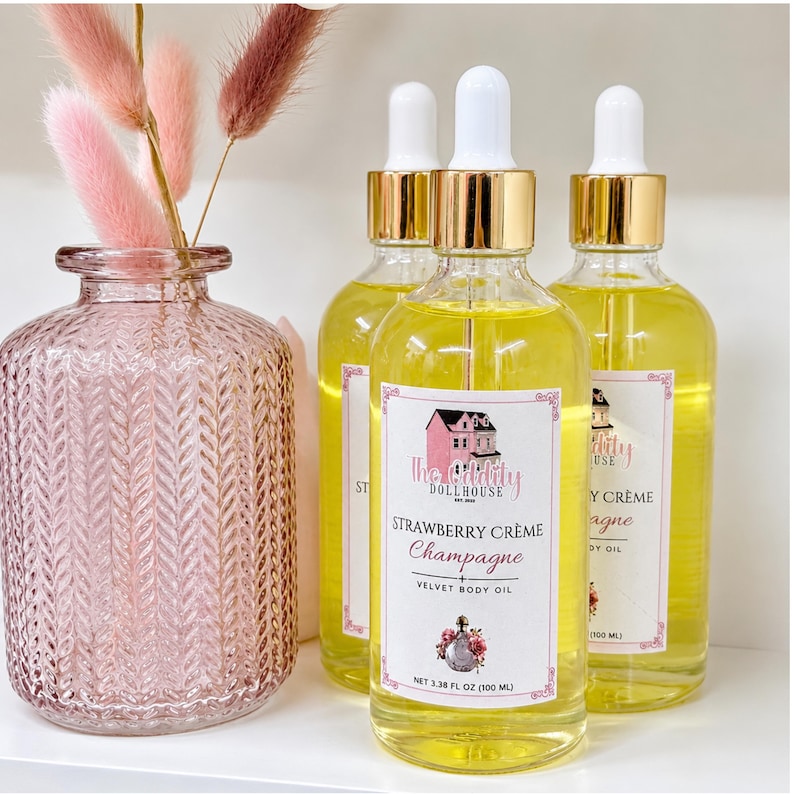 May include: Three clear glass bottles filled with yellow body oil, each featuring a white dropper cap with a gold collar. The labels read The Oddity Dollhouse Strawberry Cr&egrave;me Champagne + Velvet Body Oil, 3.38 fl oz (100 ml), with a floral graphic.