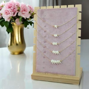 May include: A display of gold-colored chain necklaces, each featuring a white, bone-like pendant. The necklaces are arranged on a pink velvet backing within a wooden display stand. A gold vase with pink roses is in the background.