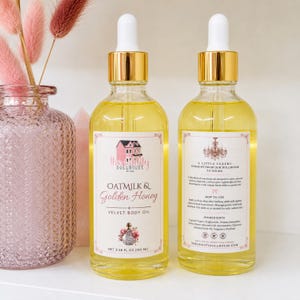 May include: Two clear glass dropper bottles filled with yellow body oil, featuring gold caps and white labels. The front label reads Oatmilk & Golden Honey Velvet Body Oil, 3.38 fl oz (100 ml). The back label includes instructions and ingredients.