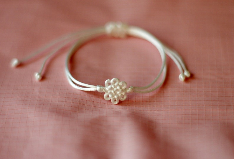 May include: A white string bracelet with a white flower-shaped knot in the center.