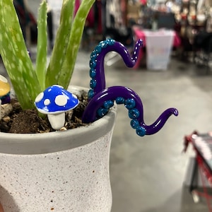 May include: A white ceramic pot with a speckled finish, containing a green aloe vera plant and a purple octopus figurine with blue accents. A white mushroom figurine with blue and white polka dots is also in the pot.