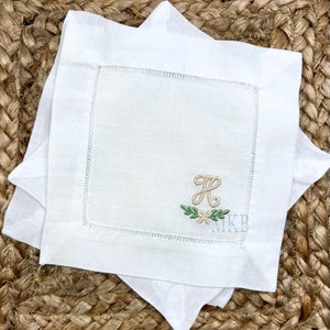 Pode incluir: Guardanapo de linho branco com uma inicial bordada "H" em fio bege com uma coroa de louro verde e flores brancas. O guardanapo tem uma borda branca costurada.