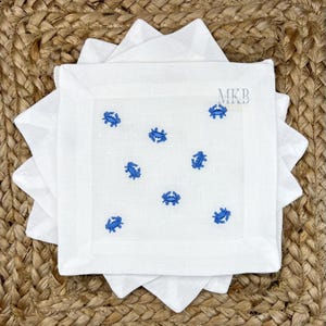 以下が含まれることがあります： 白いリネンのカクテルナプキン。青いカニの刺繍が施されています。ナプキンは正方形で、スカラップエッジがあり、積み重ねられています。MKBのロゴが隅に表示されています。