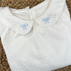 May include: White baby onesie with a peter pan collar and two blue embroidered bows on the collar. The onesie has a textured fabric.