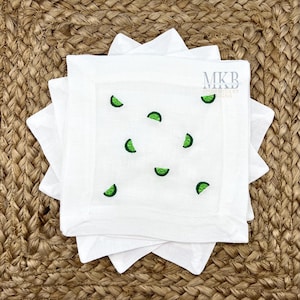 May include: A set of four white linen napkins with green embroidered lime slices. The napkins are folded and stacked on a woven brown surface. The text "MKB Embroidery Designs" is embroidered in the top right corner.