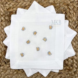 May include: White linen cocktail napkins with embroidered beer mug designs. The napkins are square and stacked, with a woven background. The embroidery features gold and silver thread, and the text "MKB Embroidery Designs".