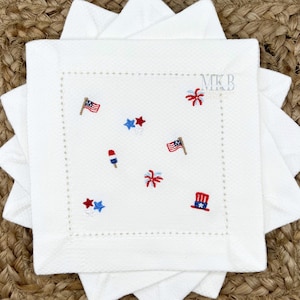 May include: White square cloth napkin with embroidered red, white, and blue patriotic designs. Features American flags, stars, fireworks, and a firecracker. The napkin has a textured surface and a stitched border.