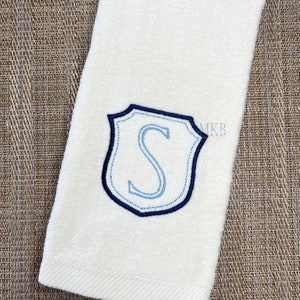 Può includere: Asciugamano bianco con uno stemma ricamato in blu che presenta la lettera "S" al centro.