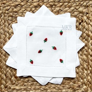 May include: White linen napkins with embroidered red strawberries and green leaves. The napkins are square with a decorative stitched border. The napkins are stacked on a woven, tan surface.