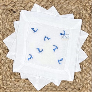 May include: Set of white linen napkins with blue embroidered outlines of the island of Martha's Vineyard. The napkins are folded and stacked on a woven surface. The text "MKB Embroidery Designs" is embroidered on one of the napkins.