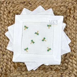 Op de afbeelding: Een stapel witte linnen servetten met geborduurde groene en gouden libellen. De servetten hebben een decoratieve gestikte rand en liggen op een geweven jute oppervlak. De servetten zijn vierkant.