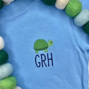 May include: A light blue baby onesie with an embroidered green turtle and the name "GRH" in navy blue.