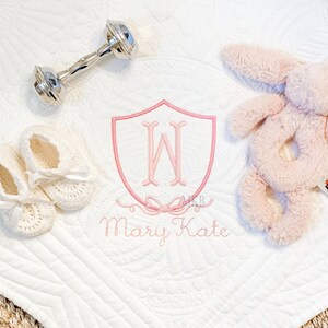 May include: A white baby blanket with a pink embroidered monogram in a shield shape. The monogram is the letter "W" with a pink bow. The text "Mary Kate" is embroidered below the monogram. The blanket is surrounded by a silver rattle, a pair of white crocheted baby shoes, and a pink plush bunny.