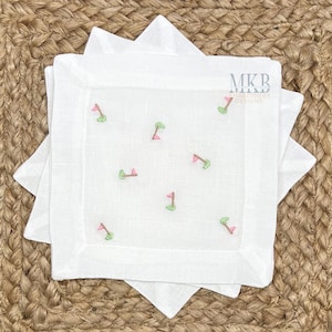 May include: Set of three white linen napkins with embroidered green golf flags and pink flags. The napkins are folded and stacked on a woven brown surface. MKB Miniature Designs is printed on the napkins.