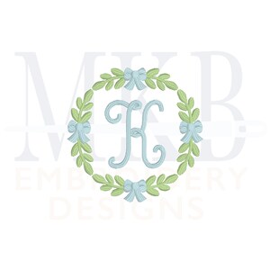 May include: A monogram embroidery design featuring the letter "K" in a light blue colour, surrounded by a wreath of green leaves and blue bows.