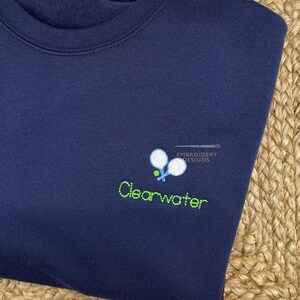 May include: A navy blue sweatshirt with a green embroidered design of two tennis rackets and a ball. The text "Clearwater" is embroidered below the rackets.