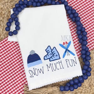 May include: White kitchen towel with blue embroidered winter-themed design. The design includes a ski hat, ski boots, skis, and the text "SNOW MUCH FUN".