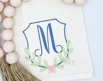 May include: A white kitchen towel with a blue and green embroidered monogram in a crest shape. The monogram is the letter "M" with a wreath of leaves and a pink bow. The towel has the initials "MKB" embroidered in the bottom right corner.