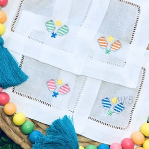 May include: Four white linen napkins with embroidered paddle ball designs. Each napkin features a different color paddle ball: green, orange, pink, and blue.