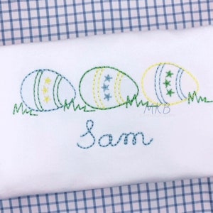 Puede incluir: Diseño bordado de tres huevos de Pascua en verde, amarillo y azul con estrellas blancas sobre una tela blanca con el nombre Sam cosido debajo.