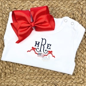 May include: White baby bodysuit with a red bow and embroidered initials. The bodysuit features a patriotic design with the American flag and red ribbons. The design includes the initials "MRe".