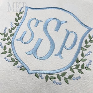 Op de afbeelding: Geborduurd monogram met de letters "SSP" in lichtblauw, geplaatst in een schildvormige lijst. Het ontwerp is omgeven door een krans van groene bladeren en kleine blauwe bessen, allemaal gestikt op een crèmekleurige stof. De initialen zijn in een elegant lettertype.