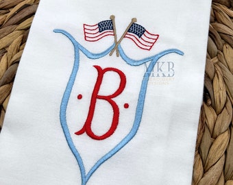 May include: An embroidered monogram featuring the letter "B" in red, surrounded by a blue and white design. Two small American flags are crossed above the monogram.