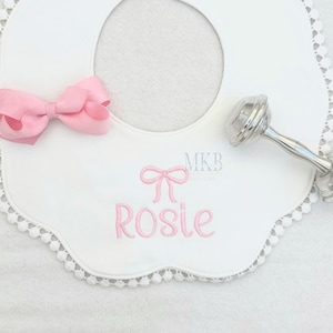 May include: White scalloped bib with a pink bow and the name "Rosie" embroidered in pink. A silver rattle is also pictured.