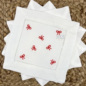 Peut inclure: Serviettes en lin blanc avec un motif de nœud rouge brodé. Les serviettes sont pliées et empilées les unes sur les autres. Le texte "MKB" est brodé sur la serviette.